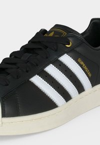 Μαύρο παπούτσι Adidas Superstar με λευκές τρεις ρίγες, μαύρα κορδόνια, χρυσά πόδια και χρυσό κείμενο "SUPERSTAR" στο πλάι.