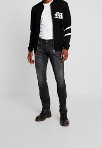 Svart zip-up kofta med vitt logotyp och ränder, tillsammans med gråjeans och svarta snörstövlar. Enkel och funktionell design.