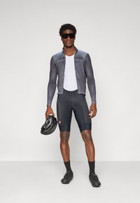 Sykkelantrekk med en elegant grå langermet trøye, sorte shorts og sorte sko. Modellen holder en svart hjelm og har på seg mørke solbriller.