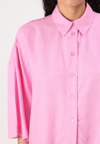 Rosa skjorte med knapper laget av lett, teksturert stoff. Har en spiss krage og seks matchende knapper nedover fronten.