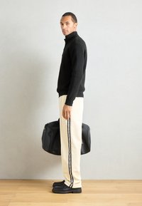 Svart strikkegenser med høy krage; kremfargede sweatpants med en svart stripe; kraftige svarte sko; holder en svart veske.