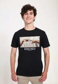 Černé bavlněné tričko s centrálním grafickým motivem rukou, které se navzájem dotýkají, s textem „MICHAELANGELO“ a „STVOŘENÍ ADAMA“. 