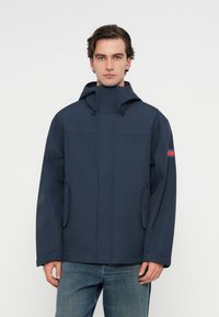 Ναυτικό μπλε αδιάβροχο μπουφάν με ψηλό γιακά, ρυθμιζόμενη κουκούλα, φερμουάρ μπροστά και πλαϊνές τσέπες; λεία υφή και μινιμαλιστικός σχεδιασμός.