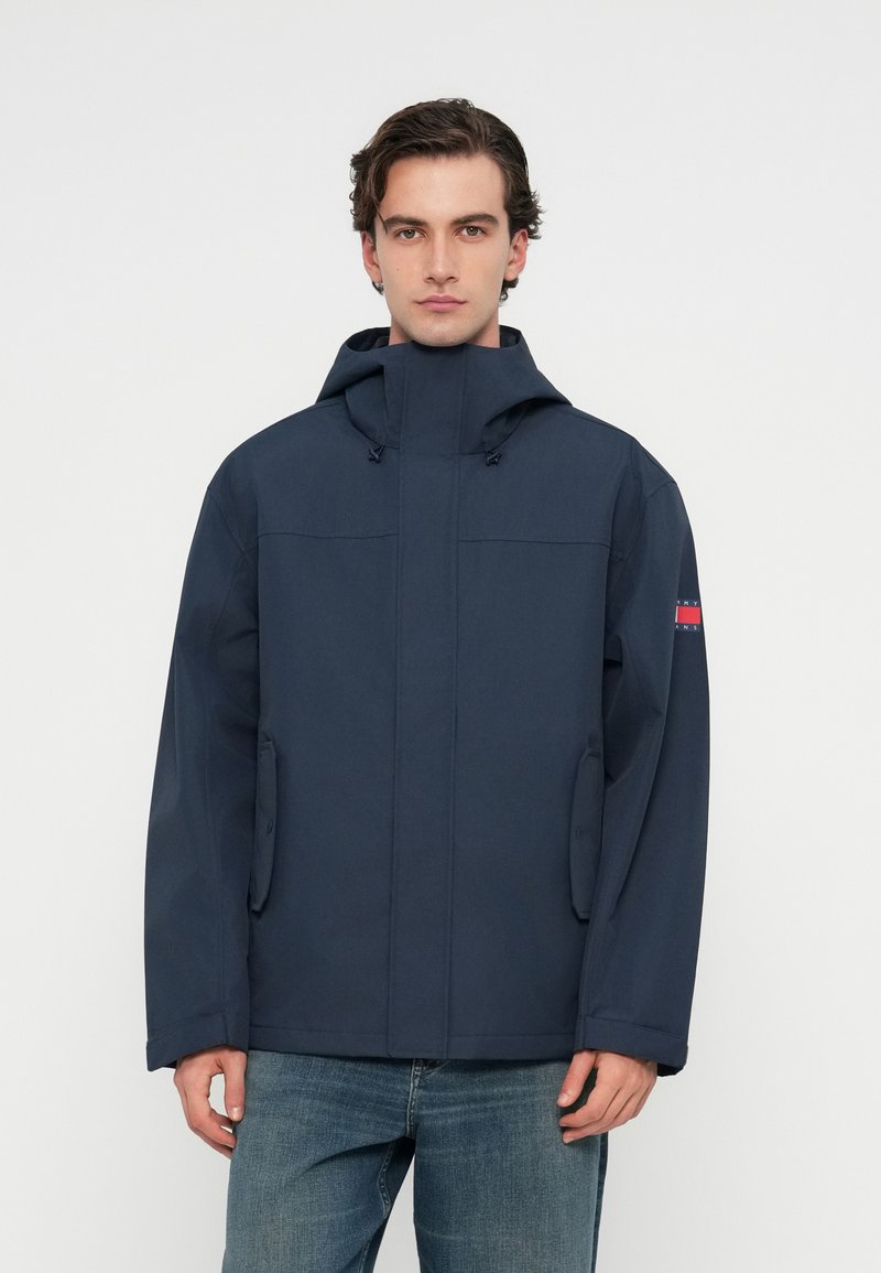 Ναυτικό μπλε αδιάβροχο μπουφάν με ψηλό γιακά, ρυθμιζόμενη κουκούλα, φερμουάρ μπροστά και πλαϊνές τσέπες; λεία υφή και μινιμαλιστικός σχεδιασμός.