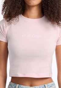 Šviesiai rožinė trumpa marškinėliai, su įsiūtu balto teksto užrašu "Guess Jeans", trumpomis rankovėmis ir prigludusia forma.