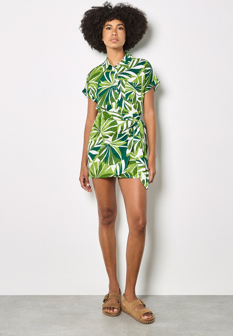 Femme aux cheveux bouclés portant une robe chemise à manches courtes à imprimé de feuilles tropicales vertes et blanches, ainsi que des sandales couleur sable, debout contre un mur blanc.