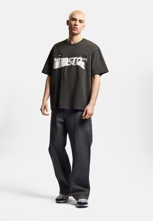 Młoda osoba z krótkimi blond włosami, ubrana w luźną czarną koszulkę z nadrukiem, szerokie ciemne jeansy i wzorzyste sneakersy, stojąca na białym tle.