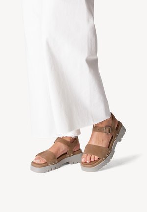 Sandali in suede marrone con una suola grigia spessa, dotati di due cinturini larghi e un cinturino alla caviglia con fibbia. Parte superiore texturizzata con borchie metalliche.