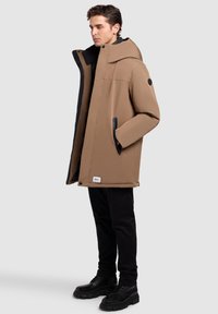 Brun isolerad parkas med huva, sidofickor och svart insida. Materialet är slätt med en matt finish. Bärs tillsammans med svarta byxor och stövlar.