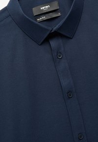 Camisa ajustada de color azul marino con una tela texturizada y un cuello clásico. Presenta botones negros y una etiqueta de cuidado en el escote.