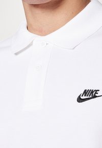 Valkoinen polo-paita, jossa on teksturoitu kangas, kaksi nappia kauluksessa ja musta brodeerattu Nike-logo vasemmassa rinnassa.