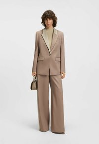 Lichtbruine op maat gemaakte suit met een beige kraag, wijde broekspijpen, twee voorzakken en een knoopsluiting. Model heeft een kleine handtas.