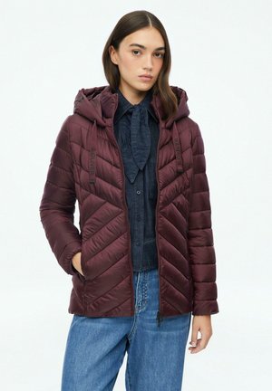 Jeune femme portant une veste matelassée à capuche rouge foncé sur une chemise boutonnée bleu foncé avec une cravate et un jean bleu, debout avec une main dans la poche.