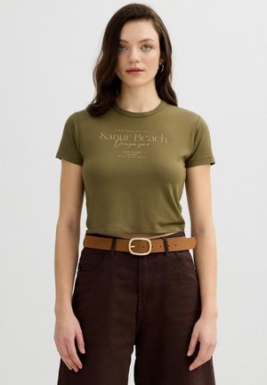 Mujer joven con cabello oscuro que lleva una camiseta corta de color verde oliva con el texto "Sanur Beach" y pantalones de talle alto de color marrón oscuro con un cinturón color canela.