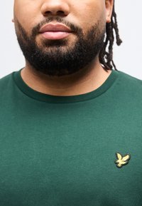 Άτομο με μουστάκι που φοράει πράσινο πουκάμισο με στρογγυλή λαιμόκοψη και ένα μικρό κίτρινο έμβλημα πουλιού στο αριστερό μέρος του στήθους, ουδέτερο φόντο.