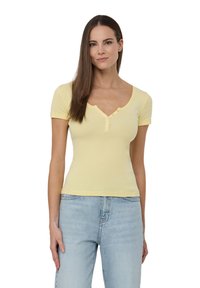 T-shirt giallo a maniche corte con scollo a V e piastra con tre bottoni, realizzato in un tessuto morbido ed elasticizzato, abbinato a jeans azzurri chiari.