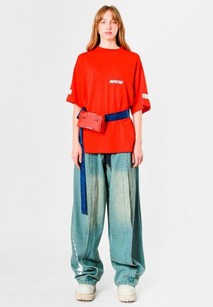 Camiseta roja de gran tamaño con logo "MWM" y bolsillo frontal, combinada con pantalones anchos de mezclilla azul claro, acentuados con una riñonera roja y un cinturón azul.
