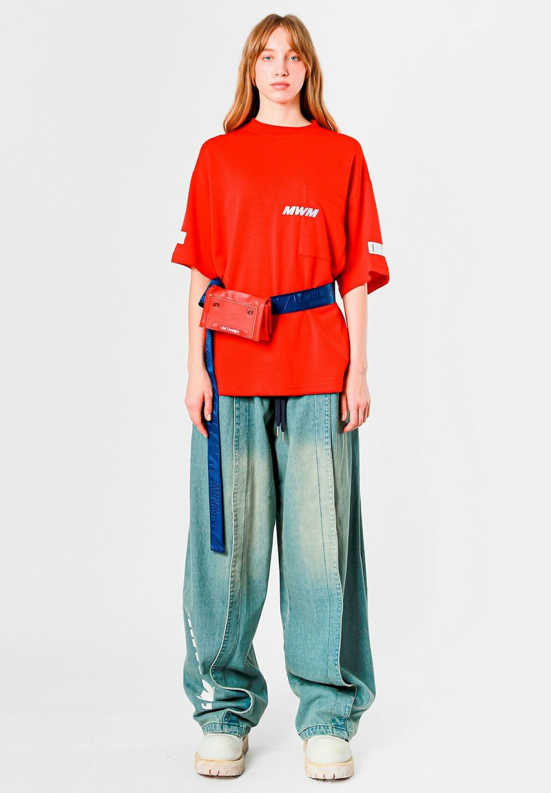 Camiseta roja de gran tamaño con logo "MWM" y bolsillo frontal, combinada con pantalones anchos de mezclilla azul claro, acentuados con una riñonera roja y un cinturón azul.