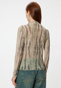 Lange mouw sheer top in beige met een golvend bruin en groen patroon, met een ronde hals en een slim fit ontwerp, gecombineerd met blauwe jeans.