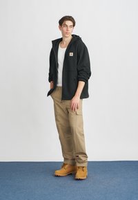 Giacca nera con cappuccio sopra una T-shirt bianca, abbinata a pantaloni cargo khaki e stivali beige, con una patch del logo sul lato sinistro.