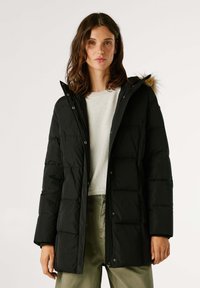 Melna pūkaina jaka ar rāvējslēdzēju un pogu aizdari, kurai ir kažokādas oderējums kapuces iekšpusē un quilted tekstūra. Valkāta virs gaismas džemperis un biksēm.