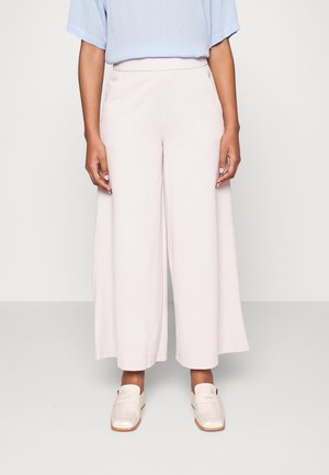 Person trägt helle rosa weite, gekürzte Hosen, hellbeige Loafers und eine hellblaue Bluse und steht vor einem einfarbigen Hintergrund.