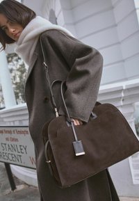 Donkerbruin suède handtas met een gestructureerd ontwerp, voorzien van twee handgrepen en een verwijderbaar etiket, afgewerkt met zwart leren hardware.