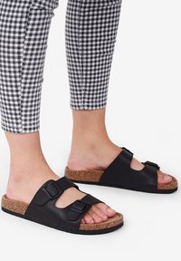 Schwarze Leder-Sandalen mit zwei verstellbaren Riemen und Korkfußbett. Flache Gummisohle; freiliegende Zehen und strukturierte Oberfläche. Gingham-Hosen sichtbar.