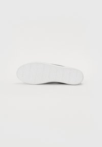 Vit sneakersula med en texturerad greppmönster, rund tå och lätt bågstöd med varumärkeslogotyp präst på sidan.