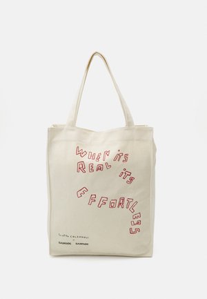 Bolsa tote beige con asas largas que presenta texto rojo que dice "When it's real it's effortless" y pequeños logos de marca negros debajo.