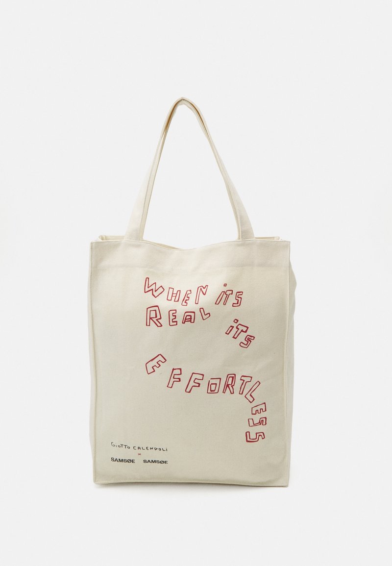 Sac fourre-tout beige avec de longues anses, comportant un texte rouge avec l'inscription "When it's real it's effortless" et de petits logos de marque noirs en dessous.