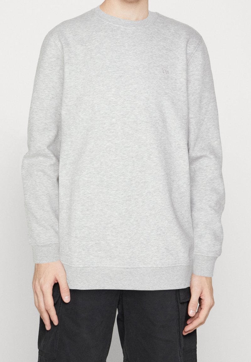 Personne portant un sweat-shirt gris clair à col rond et un pantalon noir, debout devant un fond blanc uni, visible des épaules aux cuisses.