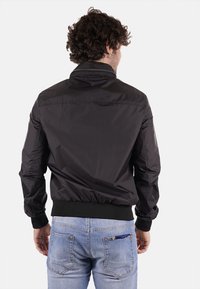 Giacca bomber in nylon nera con polsini e orlo a costine. Presenta un colletto rialzato e una texture liscia. Indossata su jeans azzurri chiari.