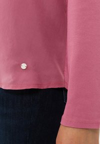 Rosa Langarmshirt mit einem runden weißen Knopfakzent am Saum. Weicher Stoff mit glatter Textur und einem leicht lockeren Sitz.
