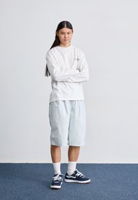 Hvit langermet skjorte, lyseblå shorts og marineblå sneakers med hvite aksenter. Modellen står med armskrysset mot en nøytral bakgrunn.