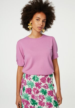 Rosa Kurzarmoberteil mit umgeschlagenen Ärmelabschlüssen, kombiniert mit einem weißen Rock mit Blumenmuster, der grüne und rosa Blätter sowie Blumen zeigt.