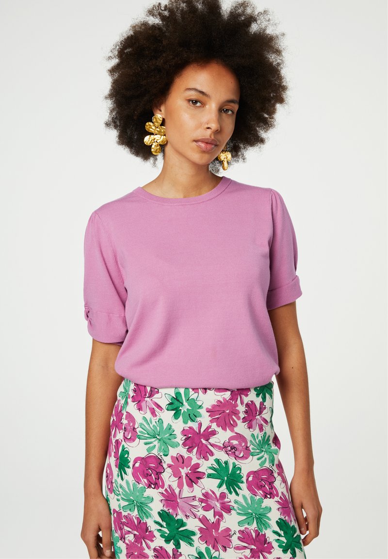 Roze T-shirt met korte mouwen en opgerolde mouwen, gecombineerd met een witte rok met bloemenprint met groene en roze bladeren en bloemen.