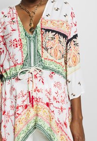 Žena v mnohobarevném květinovém tunice se zelenými, červenými, oranžovými a černými detaily a zlatým řetízkem s kruhovými přívěsky.