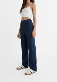 Pantalon large bleu marine en tissu lisse, avec fermeture à boutons et détails de plis à l'avant, associé à un débardeur blanc et des baskets.