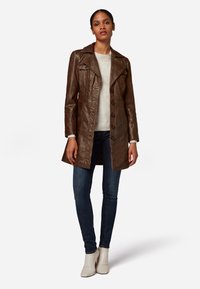 Brown leather trench coat with a wide collar, button front, side pockets, and a structured fit, worn over a light sweater and jeans.