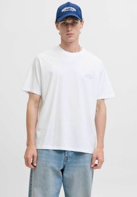 T-shirt bianca in cotone con maniche corte, scollatura a girocollo e un piccolo logo sul lato sinistro del petto. Abbinato a jeans di denim blu chiaro.