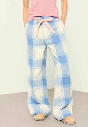 Persoon draagt losse blauw-crème geruite broek, gestrikt met een roze strik in de taille, gecombineerd met bruine instapschoenen en een roze top.