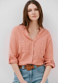 Camicia button-up di lino pesca con maniche arrotolate, caratterizzata da due tasche sul petto, infilata in jeans di denim blu chiaro, completata da una cintura marrone.