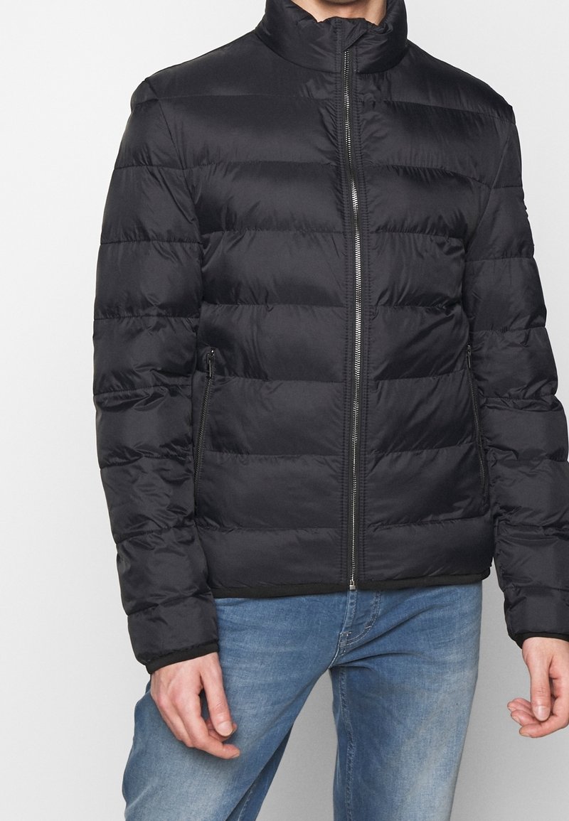 Black puffer jacket with a high collar, horizontal quilted pattern, front zipper, and side pockets, worn over blue jeans.