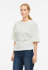 Hvit bomullstopp med korte ermer og snøring på sidene; kombinert med mørkeblå jeans. Glatt tekstur, avslappet passform.