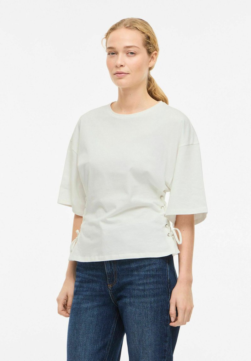 Hvit bomullstopp med korte ermer og snøring på sidene; kombinert med mørkeblå jeans. Glatt tekstur, avslappet passform.