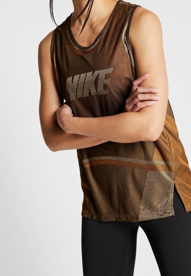 Personne portant un débardeur en mesh marron Nike, bras croisés, de la nuque aux hanches, avec un pantalon noir.