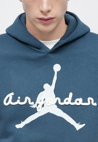 Μπλε φούτερ με κουκούλα που φέρει λευκό λογότυπο "Air Jordan" και τη σιλουέτα ενός παίκτη σε άλμα, με κεντημένες υφασμάτινες λεπτομέρειες.
