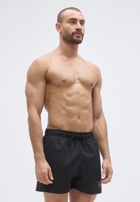 Fekete fürdőnadrág rugalmas derékrésszel és húzózsinórral. Egyszerű dizájn diszkrét márkajelzéssel. A modell izmos testalkatú.