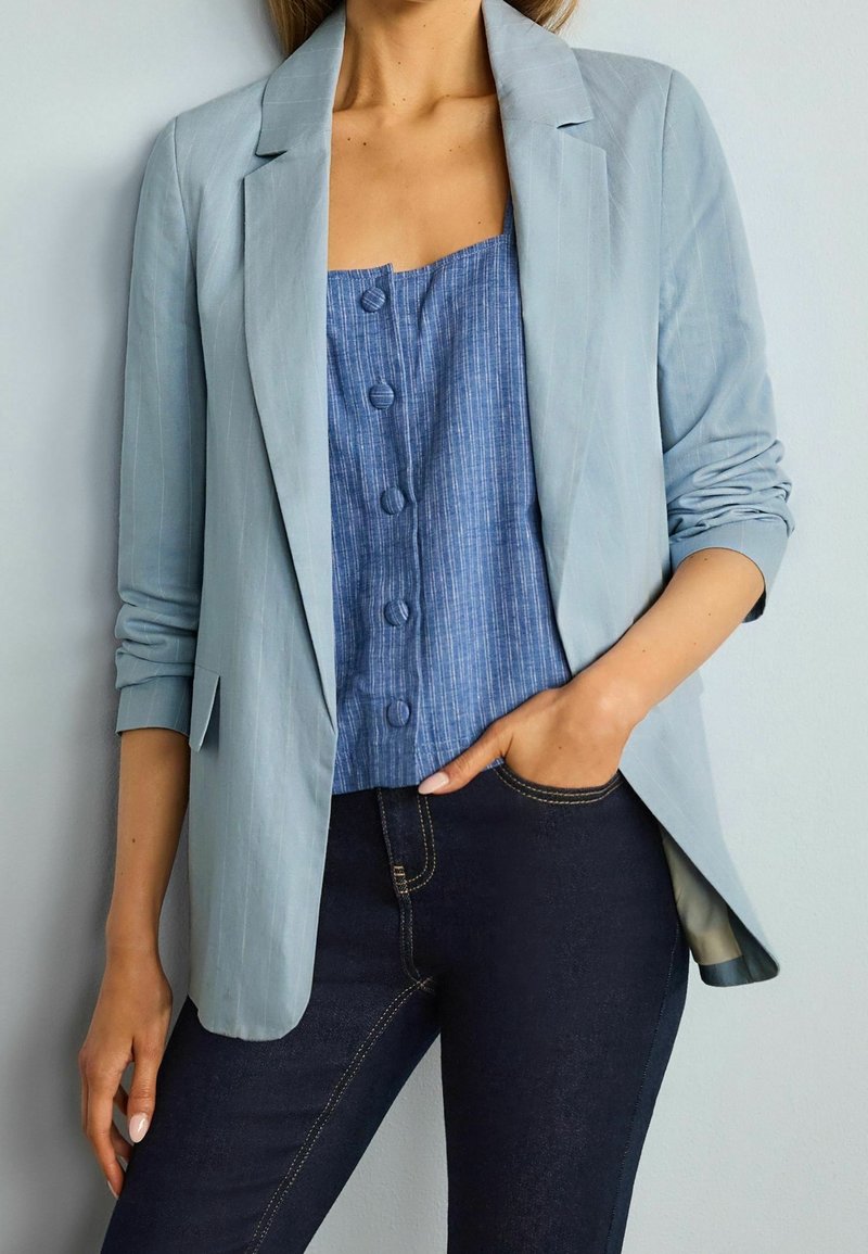 Femme portant un blazer bleu clair par-dessus un top bleu à boutons, associé à un jean bleu foncé, une main dans la poche, sur un fond uni.